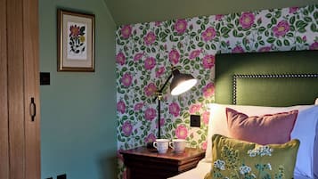 A bedroom with floral wallpaper, a bedside lamp, a framed botanical print, and a bed with pillows.