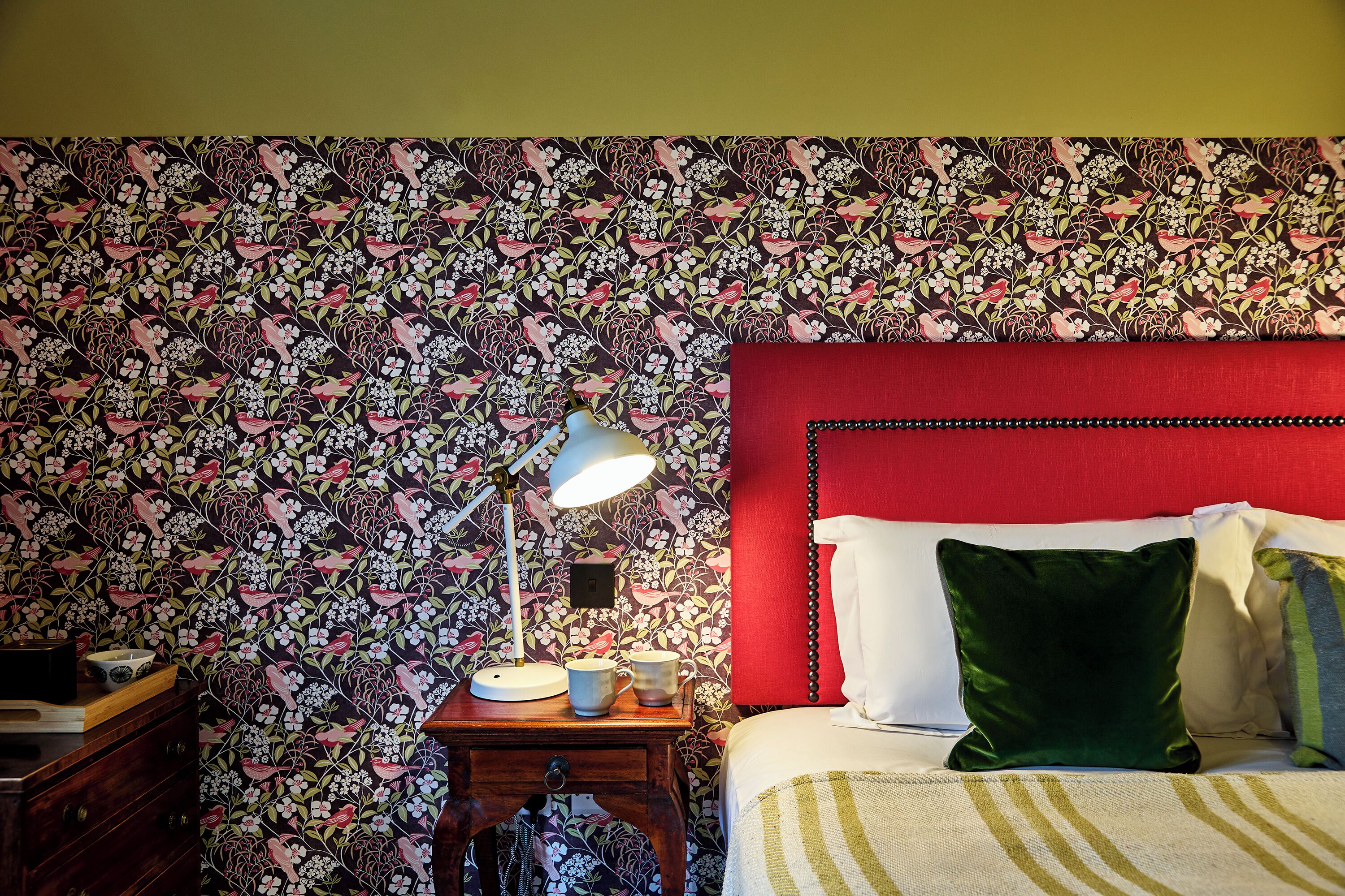 Ein Schlafzimmer mit floraler Tapete, einem roten Kopfteil und einem Nachttisch mit Lampe und zwei Tassen.