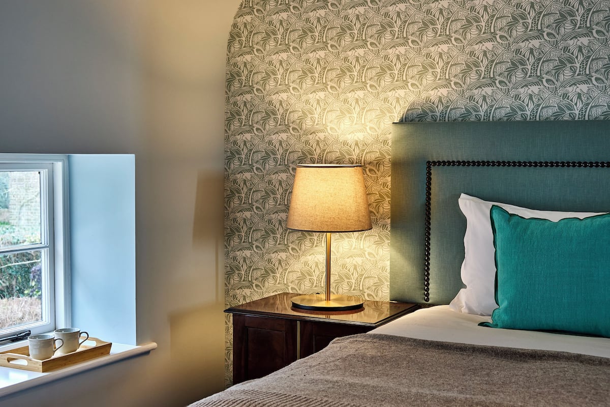 A neatly made bed with a teal pillow, a bedside lamp, and a tray with two mugs on a nightstand.