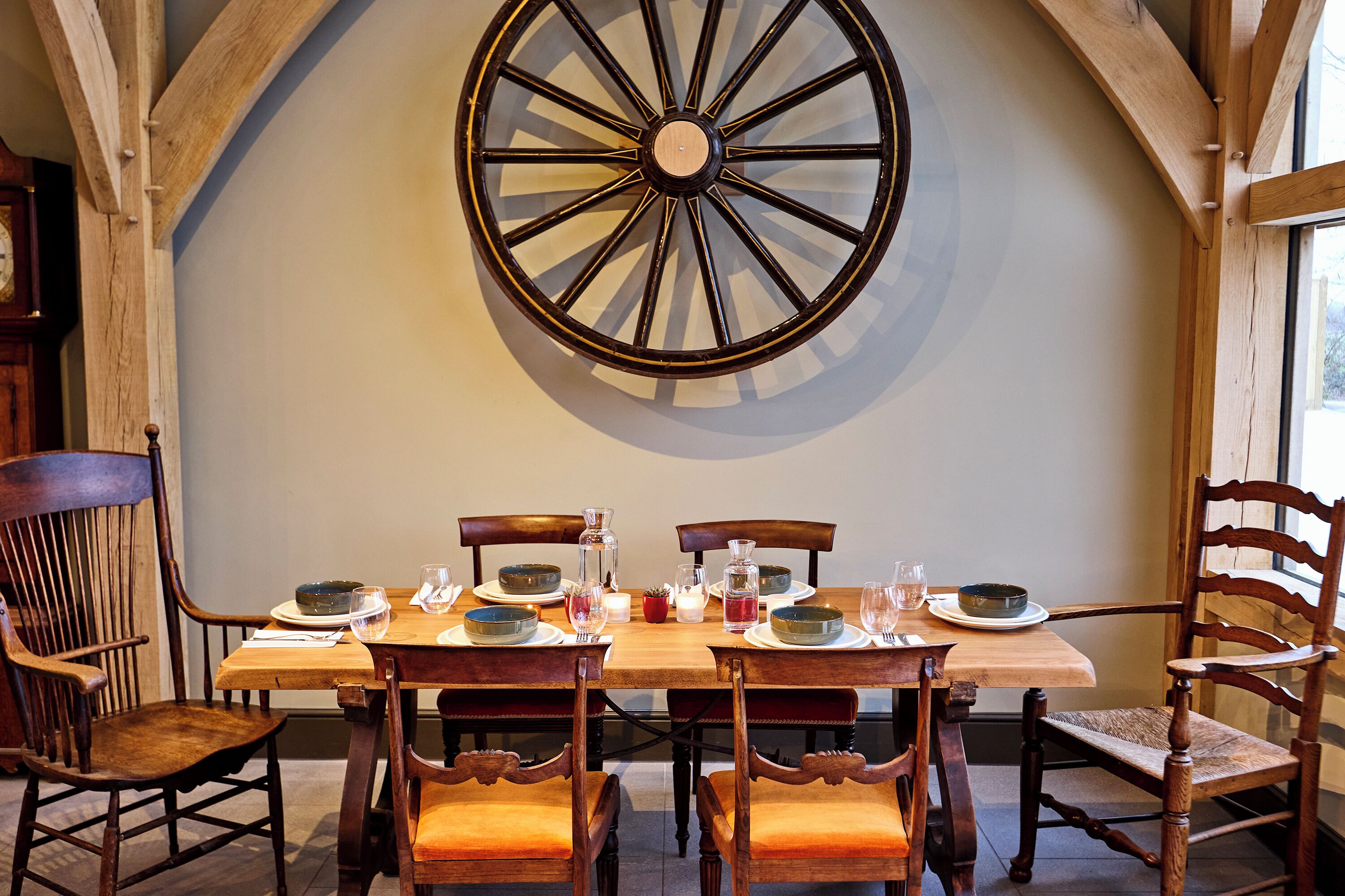 A dining area with a wooden table set for four, surrounded by wooden chairs. A large wooden wheel is mounted on the wall above the table.