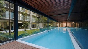 Une piscine intérieure moderne avec une terrasse en bois, des murs en verre et une vue sur un immeuble d’habitation.