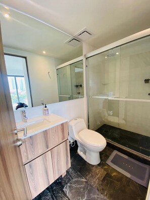 Un baño moderno con tocador de madera, inodoro blanco, cabina de ducha con mampara de vidrio y un espejo grande.