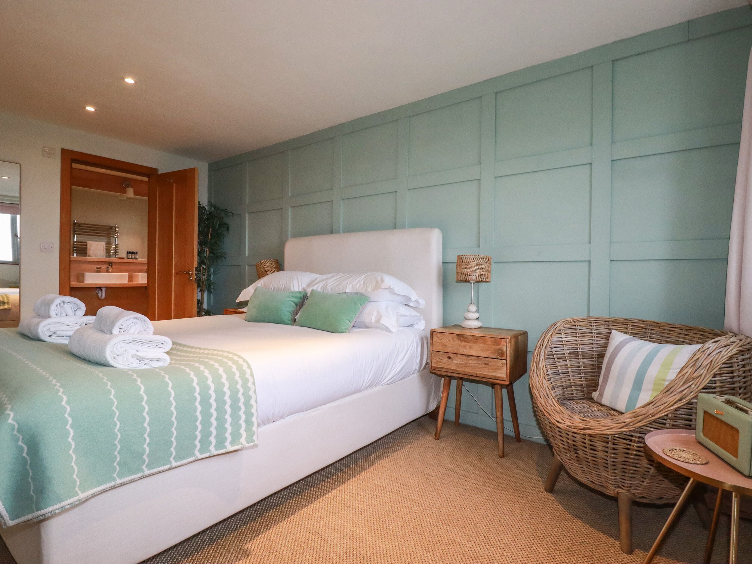 A neatly made bed with a green and white patterned bedspread, a wicker chair, a small wooden nightstand with a lamp, and a bathroom visible through an open door.