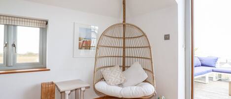 A room with a large window, a wicker chair with cushions, a small wooden table, and a framed picture on the wall.