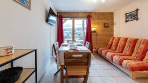 Una habitación acogedora con una mesa de comedor de madera para cuatro personas, un sofá, un televisor montado en la pared y una ventana con cortinas.