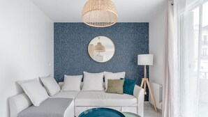 A modern living room with a white sofa, a round mirror, a blue table, and a large window with sheer curtains. - Le Rex - Bel appt avec sauna (Nantes)