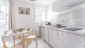 A modern kitchen with white cabinets, a dining table with chairs, a window with sheer curtains, and a butterfly painting on the wall. - Le Rex - Bel appt avec sauna (Nantes)