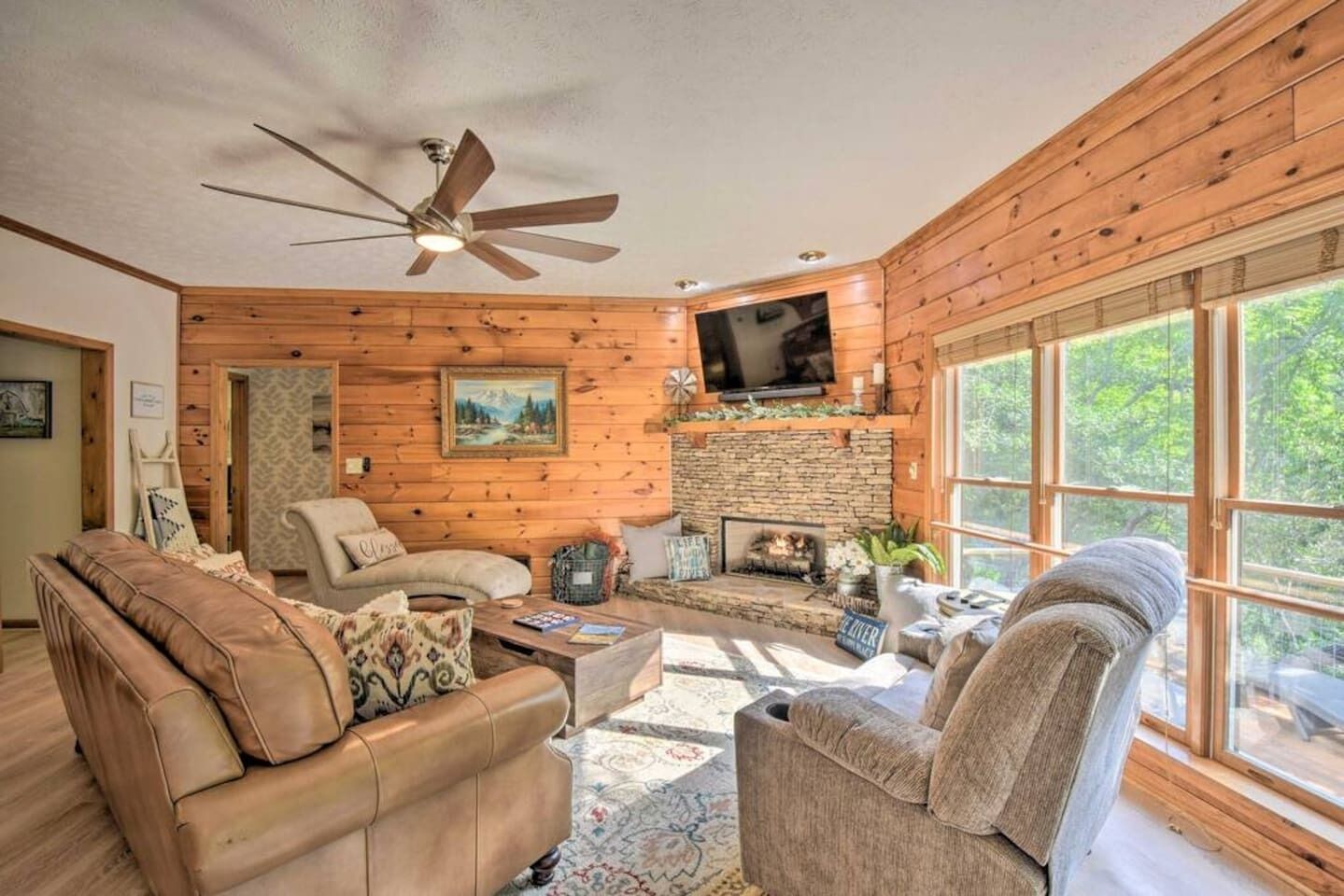 A cozy cabin interior with a fireplace, wooden walls, a ceiling fan, and a large window.