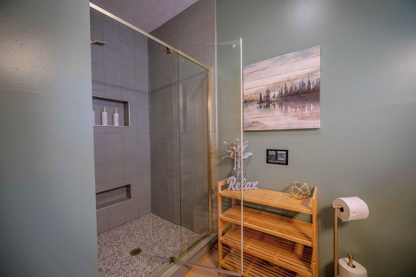 A bathroom with a glass-enclosed shower, a wooden shelf with toiletries, a framed picture on the wall, and a toilet paper holder.