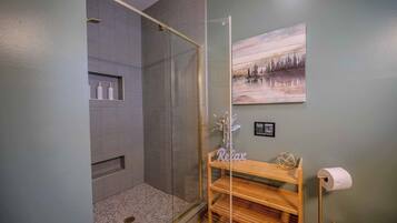 A bathroom with a glass-enclosed shower, a wooden shelf with toiletries, a framed picture on the wall, and a toilet paper holder.
