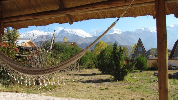 Una hamaca bajo un techo de paja, un área verde con montañas al fondo y una casa con techo triangular.