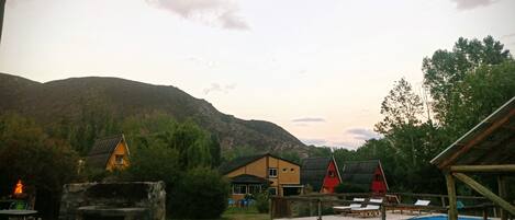 Un entorno rústico al aire libre con piscina, fogón de piedra y varias cabañas de madera.