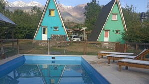 Una piscina con suelo de baldosas, dos casas turquesas con detalles de madera y un telón de fondo montañoso.