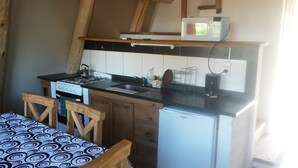 Una cocina compacta con estufa a gas, pileta, microondas y un refrigerador pequeño. Hay una silla de madera y una mesa con mantel estampado.