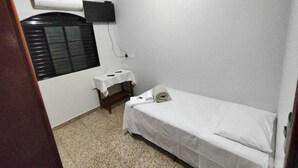 A small, clean room with a single bed, a small table with a white tablecloth, a window with blinds, and a mounted TV.