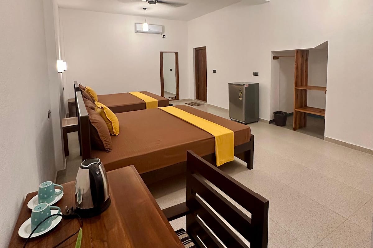 Ein Hotelzimmer mit einem großen Bett, einem Holztisch mit Wasserkocher und Tasse, einem Deckenventilator und einem kleinen Kühlschrank.