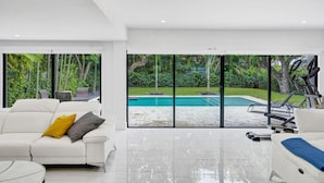 Ein modernes Wohnzimmer mit einem weißen Sofa, einem Couchtisch und einem großen Fenster mit Blick auf einen Swimmingpool und üppige Bepflanzung.