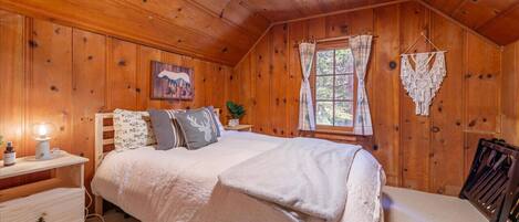 A cozy wooden cabin bedroom with a bed, nightstand, and a hanging macrame piece.