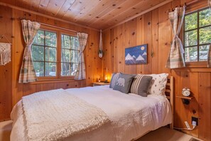 Ein gemütliches Holzblockhaus-Zimmer mit einem großen Bett, zwei mit Vorhängen versehenen Fenstern, einem Gemälde vom Nachthimmel und einem kleinen Nachttisch mit einer Lampe.