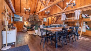 Interior rústico de una cabaña de madera con comedor, chimenea y escalera que sube a un altillo.
