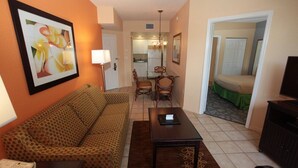 A living room with a patterned sofa, a dining table with chairs, a TV, and a kitchen area visible through an open door. - Vacation Village at Parkway. 2 bedroom, 2 bath condo that can be separate units. (Kissimmee)