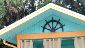 Un bâtiment turquoise orné d’un gouvernail de navire sur sa façade, doté d’un porche et de deux fauteuils de jardin recouverts d’un tissu à motifs floraux.