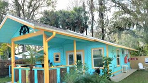 Un bâtiment aux couleurs turquoise et jaune, doté d’un porche abrité et entouré de verdure et d’arbres.