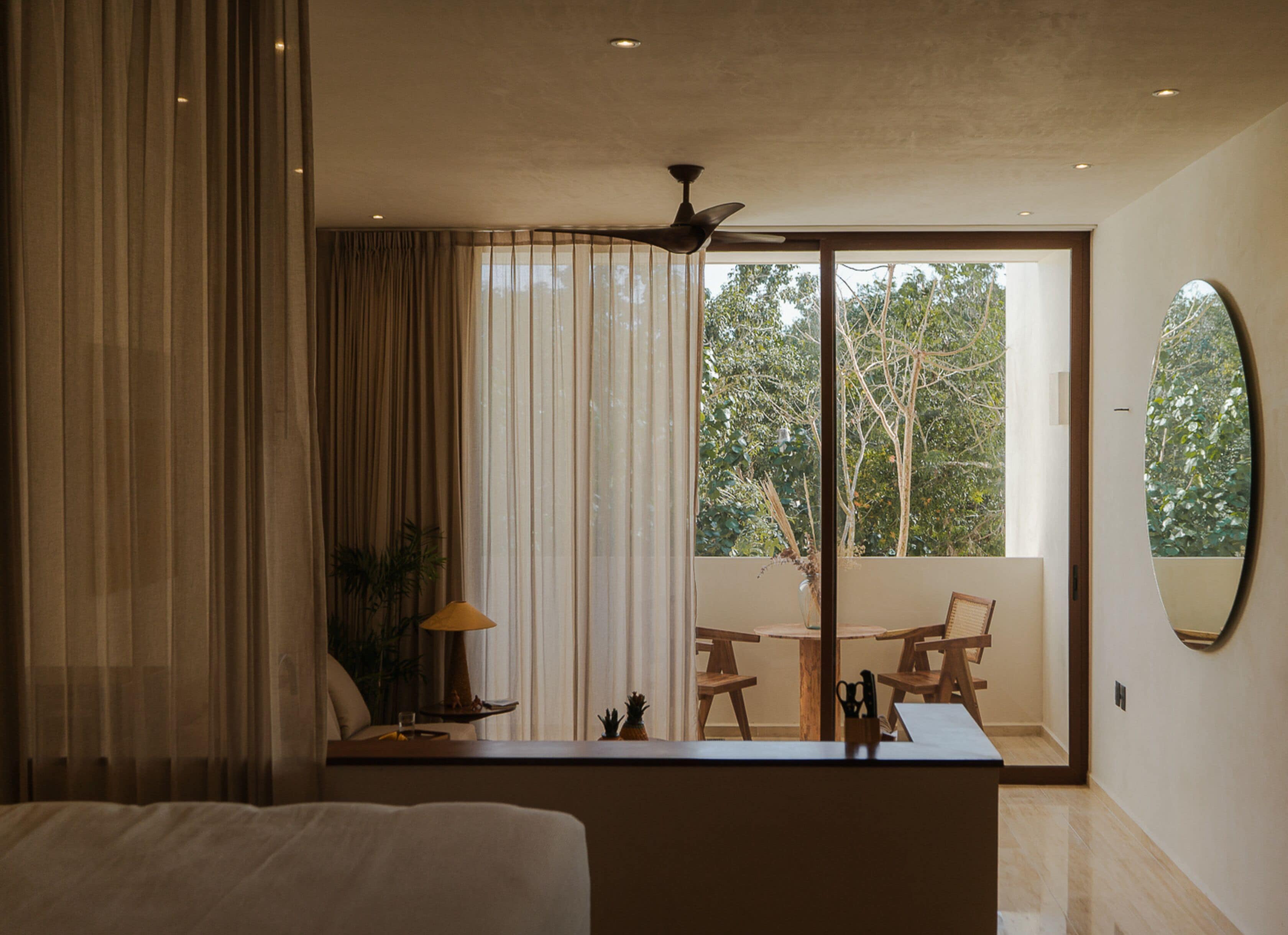 A modern bedroom with a large bed, a desk with a lamp, and a view of the outdoors through a sliding glass door.