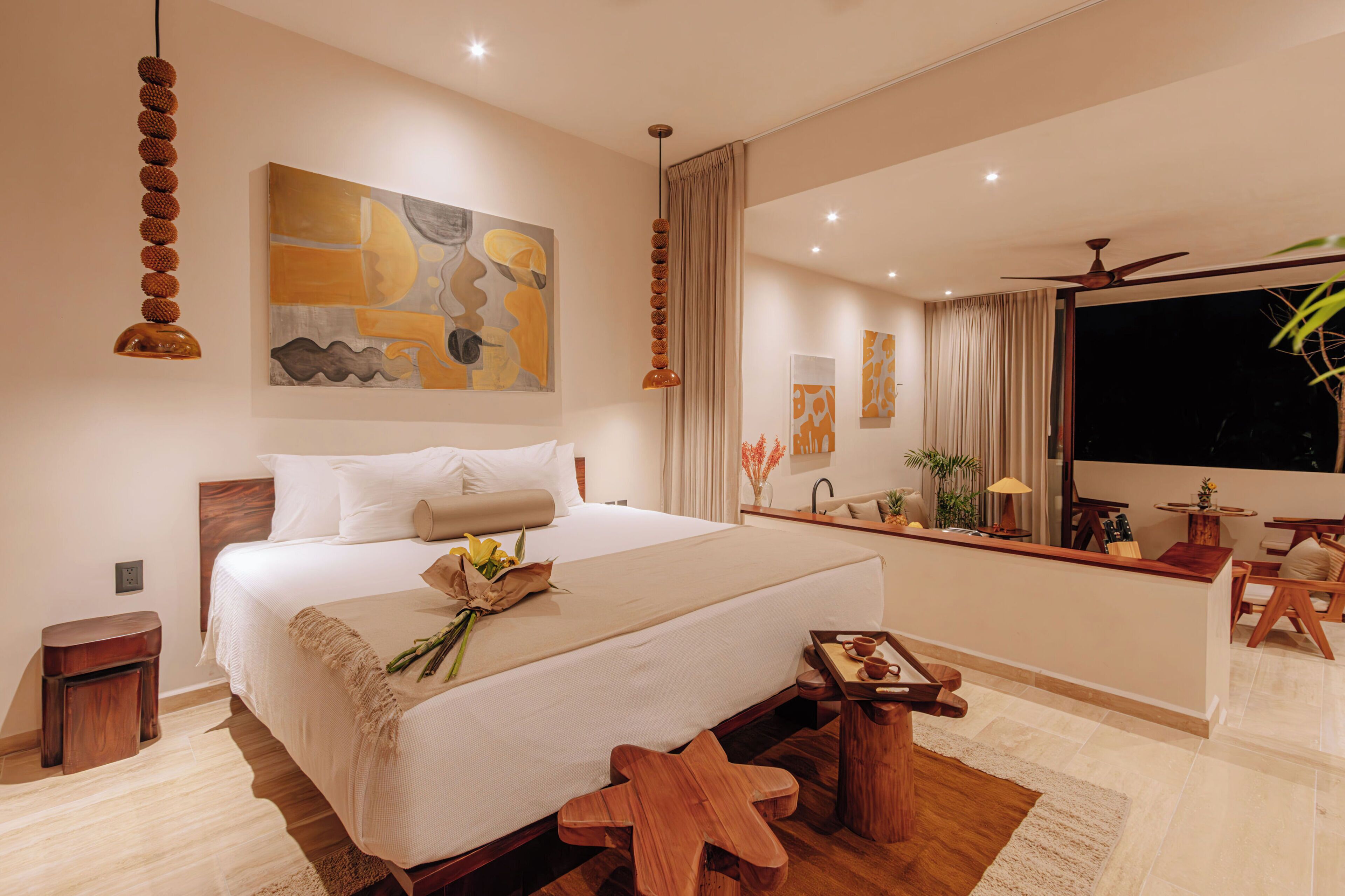 A modern hotel room with a large bed, a wooden side table, a small wooden stool, a ceiling fan, and a large window with curtains.