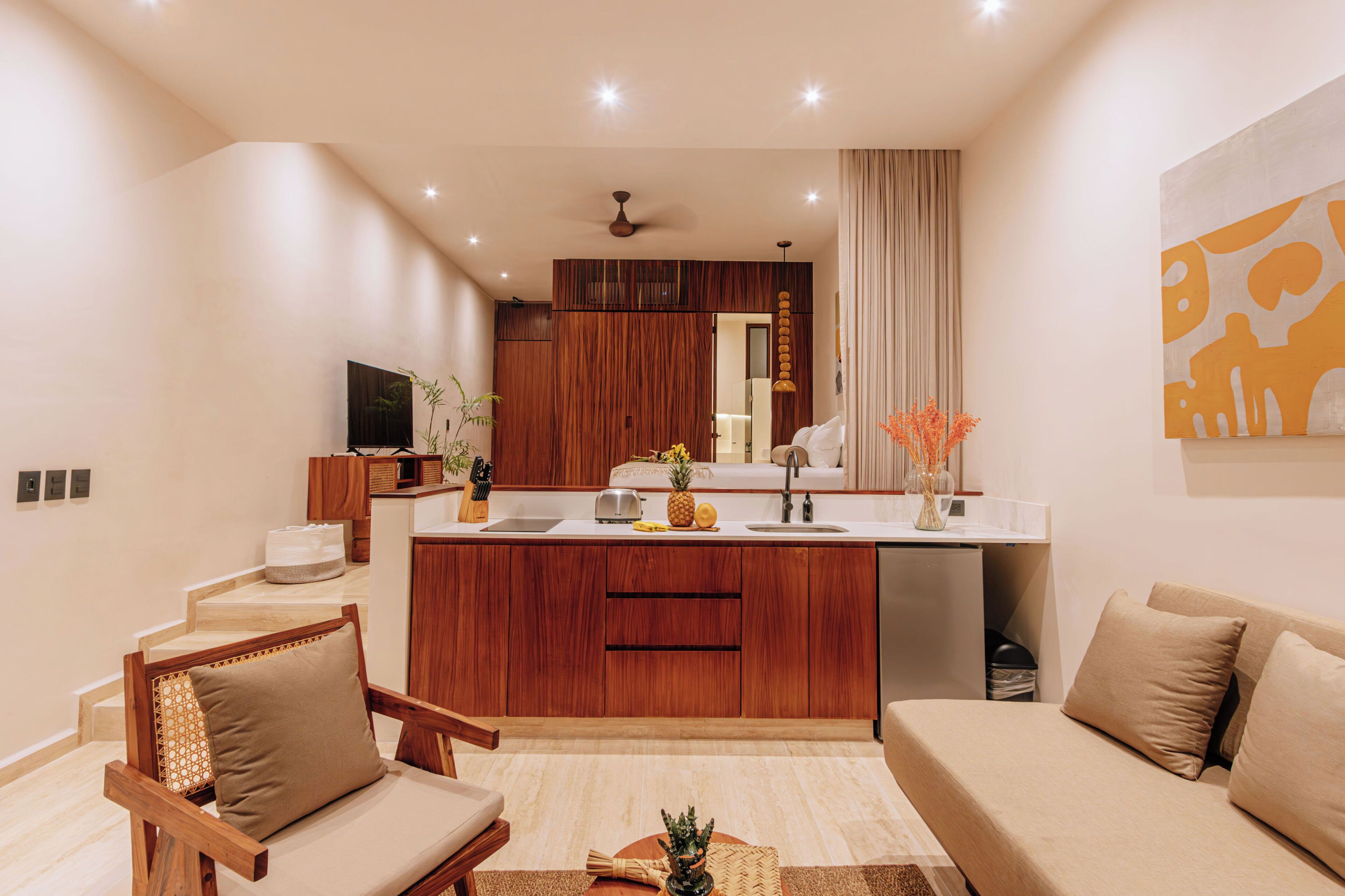Una sala moderna con un sofá, una silla de madera, una cocina con isla central y un televisor sobre un mueble.