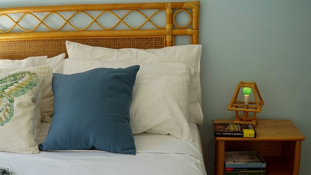 Een net opgemaakt bed met witte en blauwe kussens, een houten hoofdbord en een nachtkastje met boeken en een lamp.