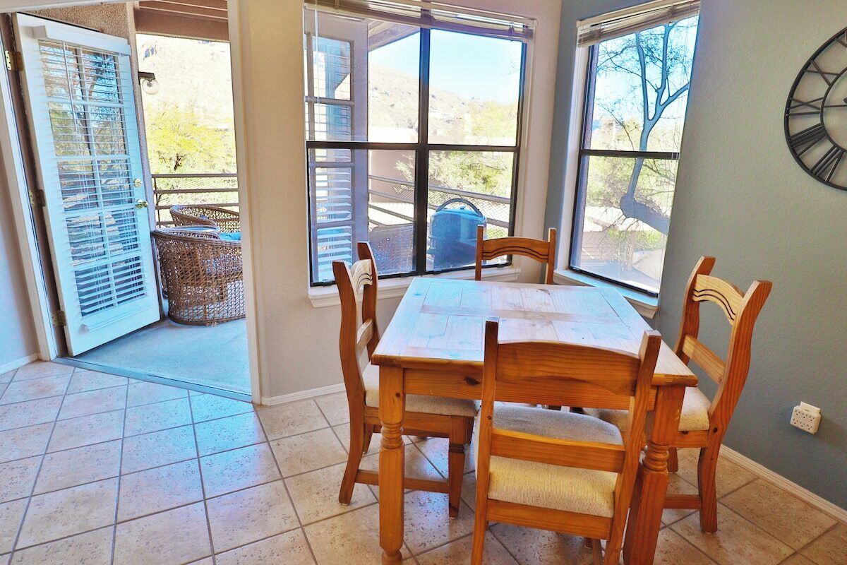 A dining area with a wooden table and chairs, a window with a view of the outdoors, and a clock on the wall.