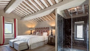 A bedroom with a bed, bedside tables, a nightstand, a window with curtains, a patterned headboard, a glass shower enclosure, and a wooden ceiling. - Villa Serena - Private villa with pool in Umbria (Umbertide)