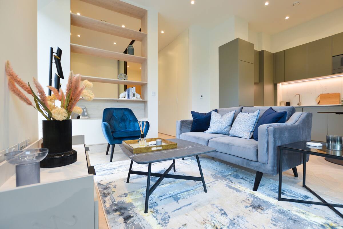Una sala de estar moderna con un sofá gris, un sillón azul y una mesa de centro. Se observan estanterías con objetos decorativos y un jarrón con flores.