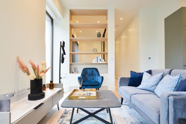 Un salon moderne avec un canapé gris, un fauteuil bleu et une table basse sur laquelle se trouve un plateau. On y trouve également une bibliothèque remplie de divers objets et une fenêtre avec un rideau.