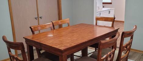 Comedor con mesa y sillas de madera, alfombra estampada y una cocina visible a través de un arco.