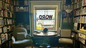Un salón de lectura acogedor con un amplio ventanal que muestra la palabra "WORLD" en el centro. Hay dos sillones, una mesita y una estantería llena de libros.