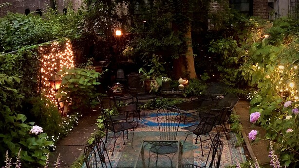Una mesa de vidrio rodeada de sillas en un área de jardín con plantas en macetas y un muro de ladrillo al fondo.