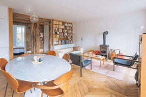 A spacious living room with a round marble table, wooden chairs, a bookshelf, a fireplace, and a large window.