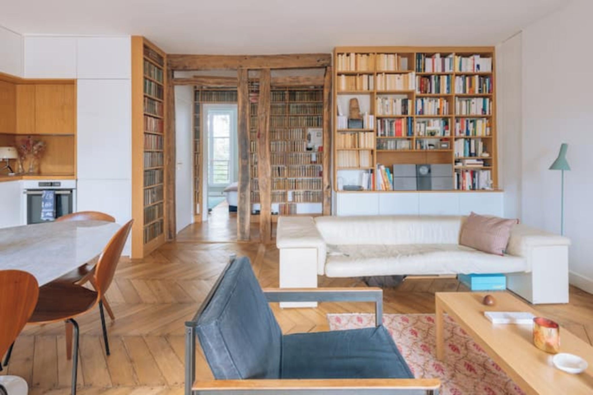 Ein großzügiger Wohnbereich mit Holzboden, einem weißen Sofa, einem blauen Sessel, einem Esstisch mit Stühlen und einem großen Bücherregal voller Bücher.