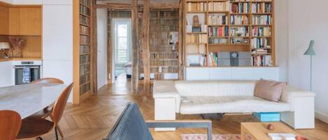 Ein großzügiger Wohnbereich mit Holzboden, einem weißen Sofa, einem blauen Sessel, einem Esstisch mit Stühlen und einem großen Bücherregal voller Bücher.