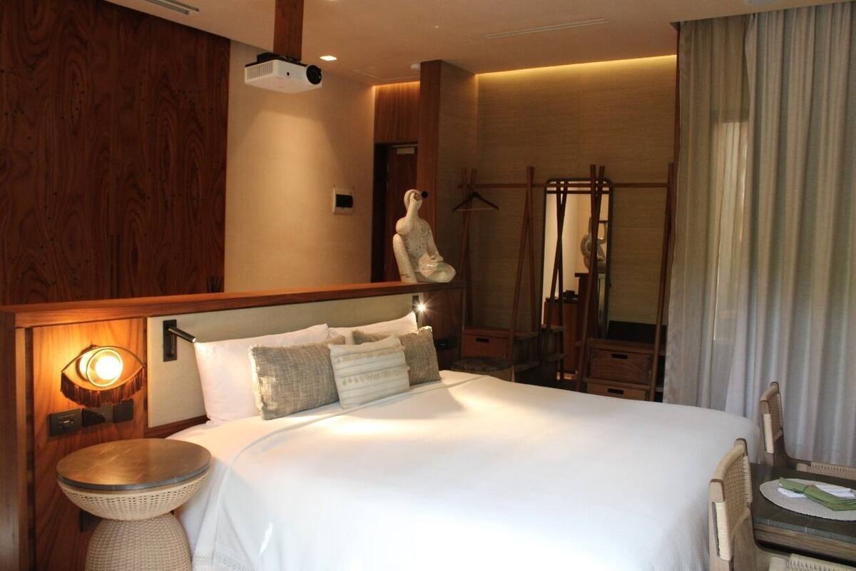 A hotel room with a large bed, a bedside table, a lamp, a wooden headboard, and a view of a wooden structure outside.