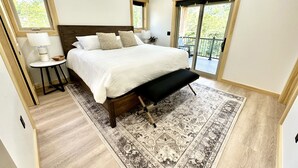 Une chambre avec un grand lit, une table de chevet, un banc, un tapis à motifs et un balcon donnant sur un paysage verdoyant.