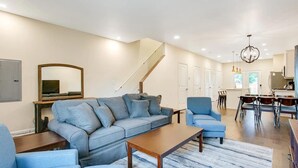 Una sala de estar amplia con un sofá modular, una mesa de centro y un comedor con sillas y una mesa. Se ve una escalera al fondo que sube a otro nivel.