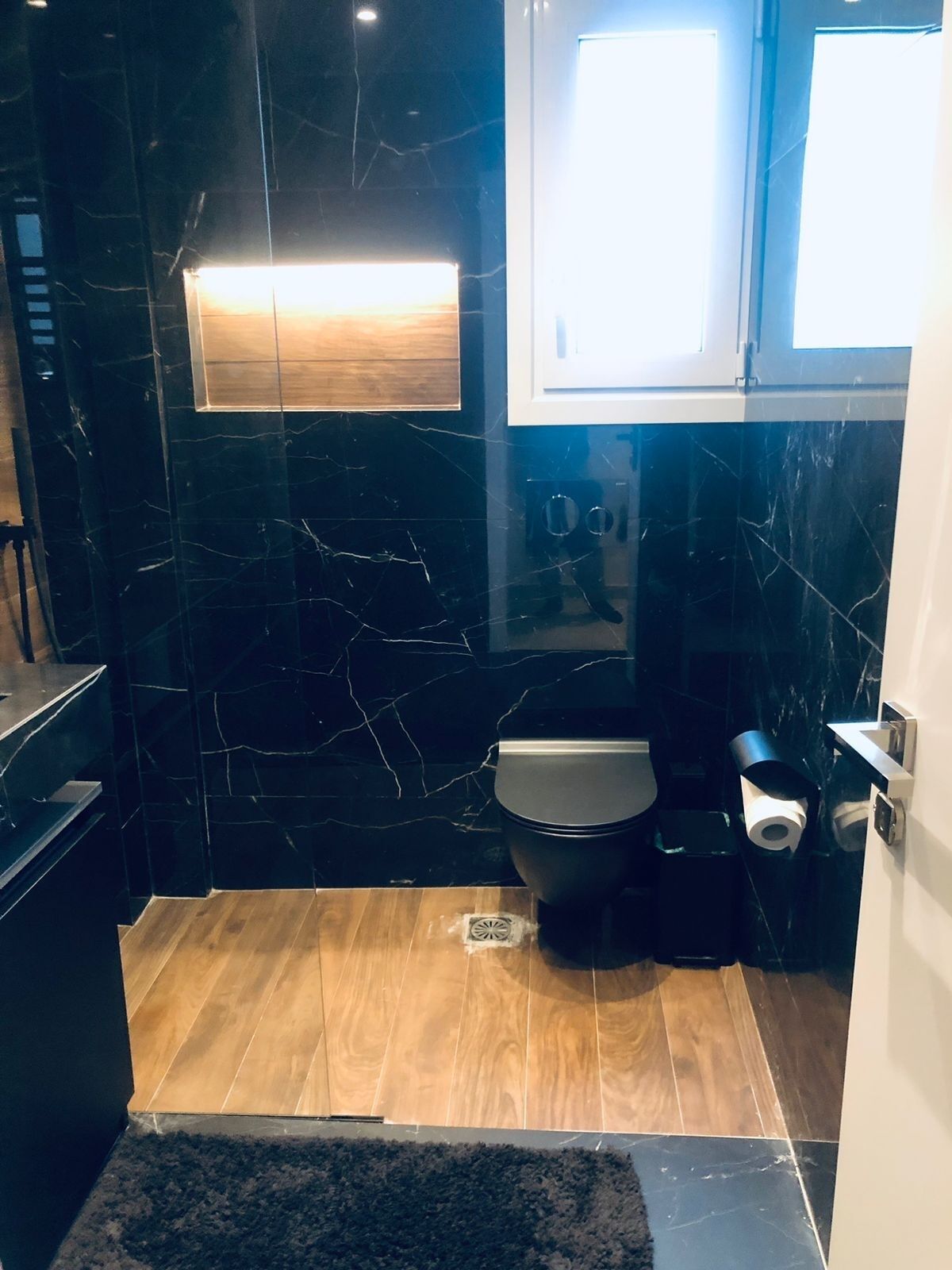 Un baño con pared de mármol negro, suelo de madera, inodoro y lavabo.
