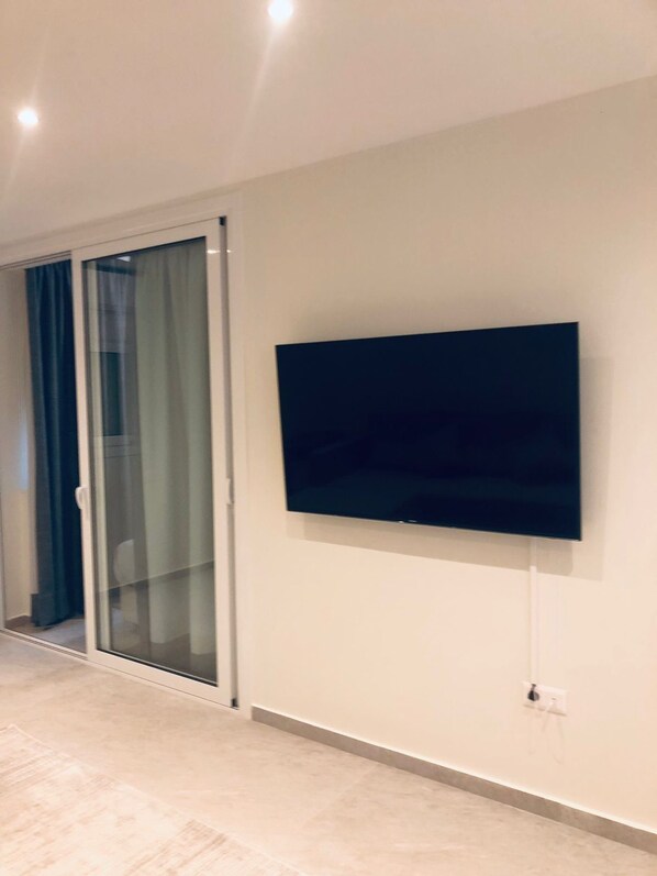 A room with a large flat-screen TV mounted on the wall, a sliding glass door with curtains, and a light-colored floor. - SeaRenity Glyfada Ctr, 1BR, Renovated, near beach (Glifada)
