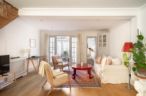 A spacious living room with a wooden floor, a red round table, a sofa, and a TV on a stand. - CHELSEA 2 BEDROOM LUXURY TOWNHOUSE - COSY AND CHARMING - WEST CENTRAL LONDON (London)
