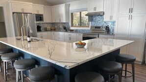 A modern kitchen with white cabinets, a large marble island with bar stools, stainless steel appliances, and a view of the outdoors. - The Perfect Place: Close to town, with 100' of private lake frontage. (Montague)