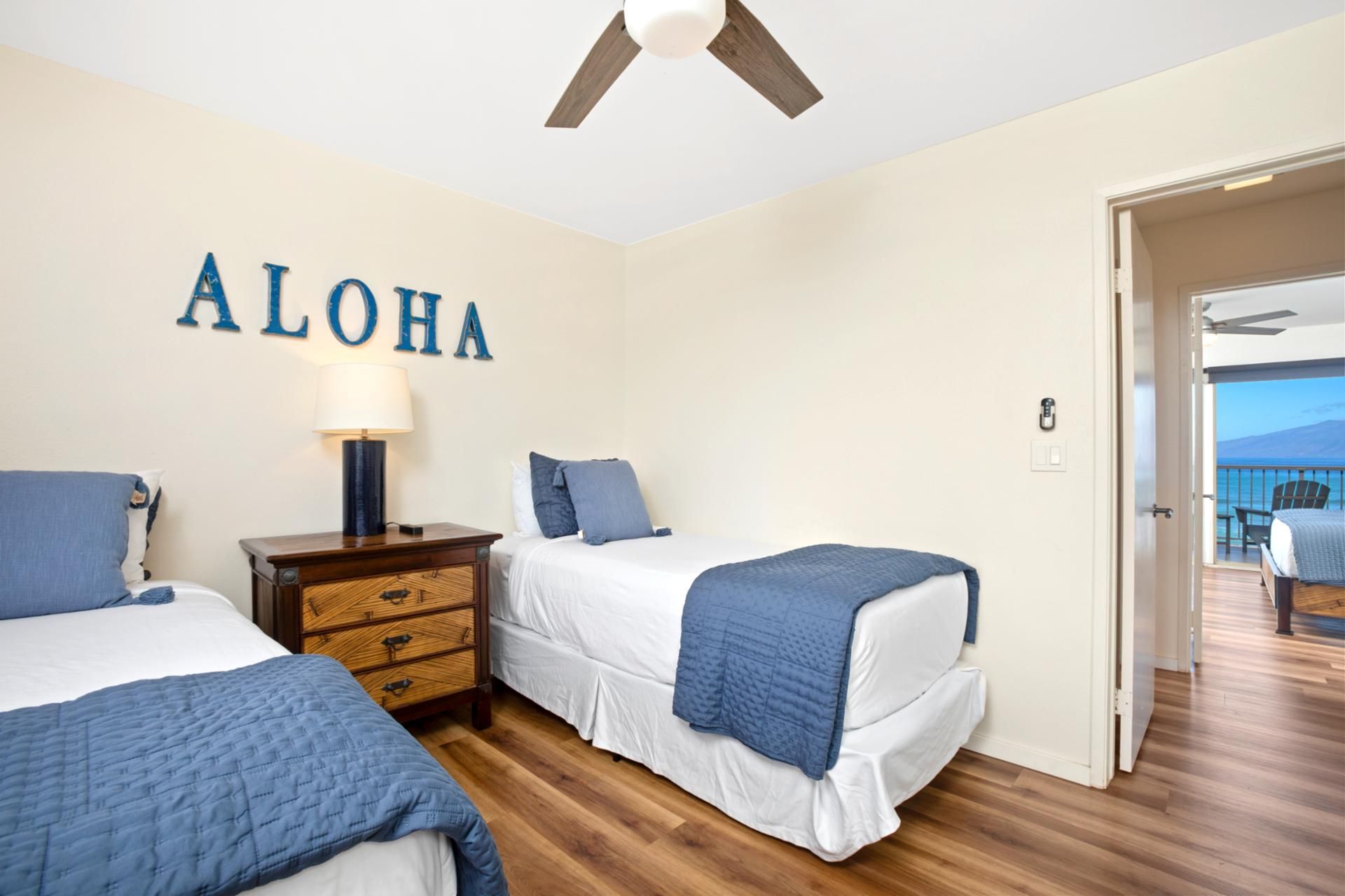 Une chambre avec deux lits, une table de chevet, un ventilateur de plafond et un mur sur lequel est écrit le mot « ALOHA ».