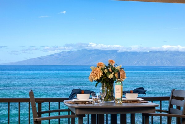 A table set with a vase of flowers, a bottle, and glasses, overlooking a body of water with mountains in the background.